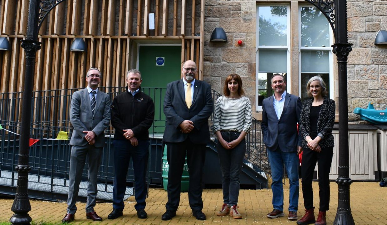 - Depute Council Leader Calum Smith standing in the centre of the image outside the rear of the newly renovated Brookwood Villa, a two storey blonde sandstone building, flanked by project partners. The group is standing underneath the magnificent portico which was saved and restored during the renovation.